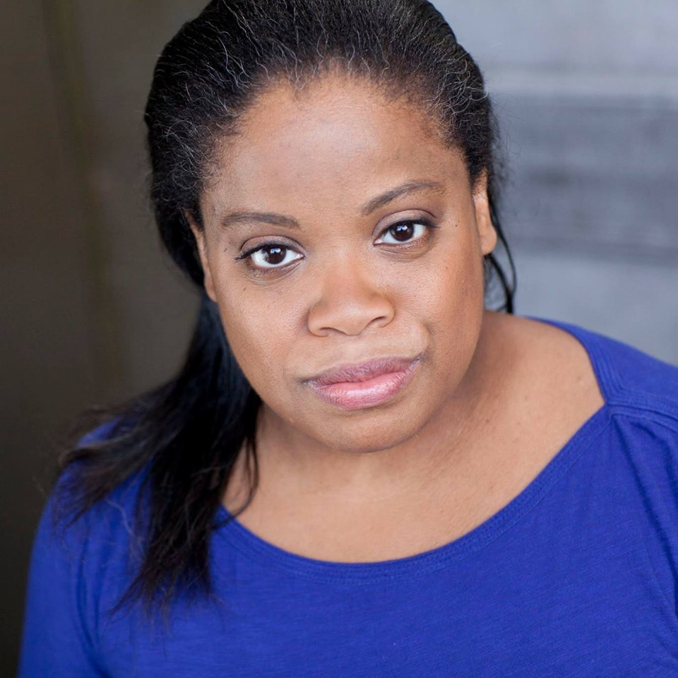 Headshot ID: A brown skinned Black woman with shoulderlength straight dark hair, greying around the edges and swept over one shoulder, looks straight into the camera with a mostly serious expression and slight smile. She is wearing an indigo shirt.
